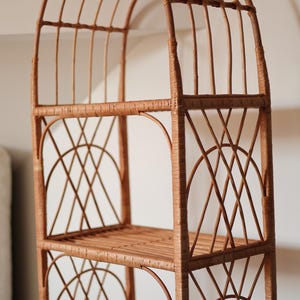 May include: A tall, freestanding wicker shelf unit with an arched top and three shelves. The unit is constructed from woven, light brown wicker, featuring a lattice design on the sides and a slatted base for each shelf. The overall design is airy and decorative.