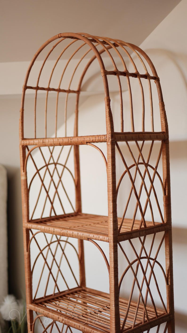 May include: A tall, freestanding wicker shelf unit with an arched top and three shelves. The unit is constructed from woven, light brown wicker, featuring a lattice design on the sides and a slatted base for each shelf. The overall design is airy and decorative.