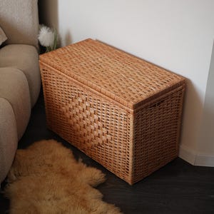 Pode incluir: Um ba&uacute; de armazenamento retangular de vime em um tom marrom quente. O ba&uacute; tem um design tecido e uma tampa articulada. &Eacute; colocado ao lado de uma parede e de um sof&aacute; bege, com um tapete de pele de ovelha na frente.