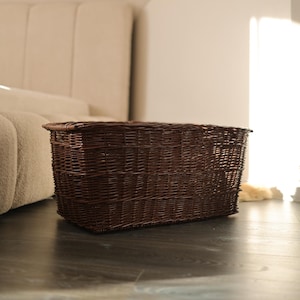 May include: A rectangular, dark brown wicker basket. The basket is made of woven material and has a sturdy, open design. It is placed on a dark wooden floor, with a neutral-colored sofa in the background.