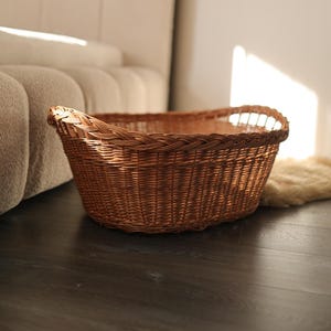 May include: A brown wicker basket with a braided edge. The oval basket is on a dark wooden floor. The basket has two handles and is empty. The basket is near a beige sofa. The basket is made of woven material.