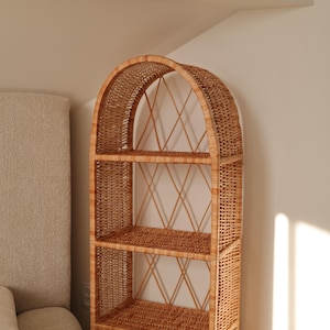 Puede incluir: Una estantería alta de mimbre arqueada con tres estantes. La estructura tejida marrón natural presenta un patrón de diamantes en la parte posterior y se coloca contra una pared de color neutro. Los estantes son ideales para exhibir libros, plantas o artículos decorativos.