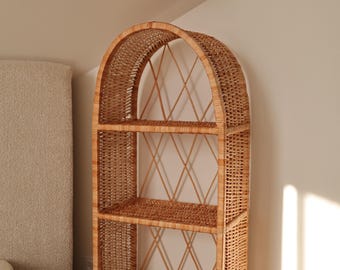 May include: A tall, arched wicker bookshelf with three shelves. The natural brown woven structure features a diamond pattern on the back and is placed against a neutral-colored wall. The shelves are ideal for displaying books, plants, or decorative items.