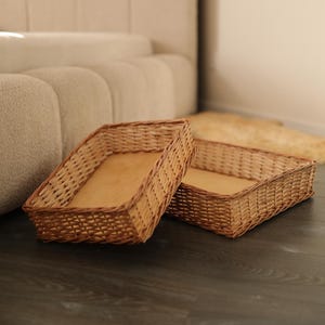 May include: Two rectangular woven wicker baskets with light brown wooden bottoms. The baskets are placed on a dark wood-grain floor. The baskets are empty and ready to be filled. The background includes a beige sofa and a light-colored rug.