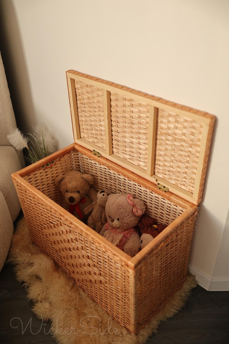 May include: A rectangular wicker storage chest with a hinged lid, filled with teddy bears. The chest is light brown and sits on a fluffy, light-colored rug. The lid is partially open, revealing the contents.