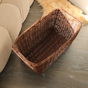 May include: A rectangular, woven wicker basket in a warm brown hue. The basket has a sturdy construction with a rectangular shape and integrated handles. It sits on a wooden floor, suggesting its use for storage or display.