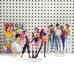 Peut inclure: Plusieurs figurines en acrylique représentant des personnages de style anime. Les personnages portent des tenues colorées, notamment du jaune, du rose et du noir. Le fond est une surface blanche avec une grille de points noirs.