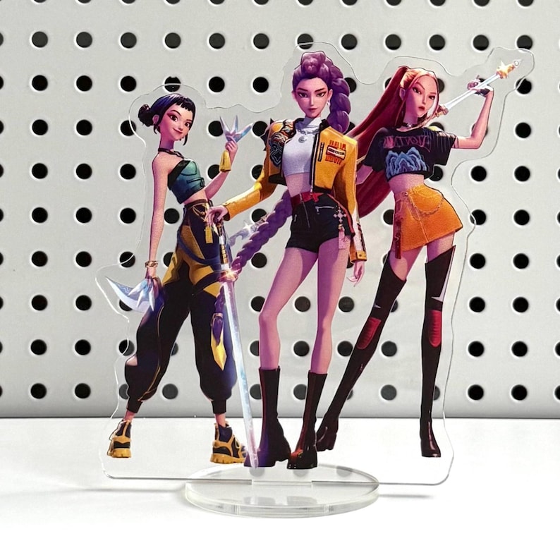 May include: A clear acrylic stand featuring three stylized female figures. The figures are dressed in fashionable outfits with vibrant colors, including yellow, black, orange, and purple. Each figure holds a weapon, and the stand has a circular base.