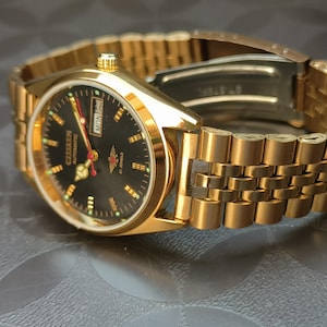 Op de afbeelding: Een goudkleurig Citizen automatisch polshorloge met een zwarte wijzerplaat, goudkleurige uurmarkeringen, een dag-datumvenster op de drie-uurpositie, een rode secondewijzer en een metalen schakelarmband met een vouwsluiting.
