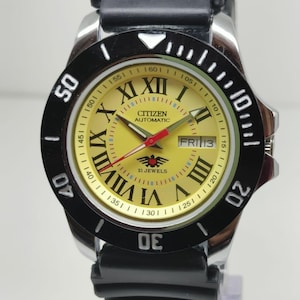 Peut inclure: Une montre-bracelet automatique Citizen avec un cadran jaune, une lunette noire et un bracelet en caoutchouc noir. La montre présente des chiffres romains, un affichage de la date et une aiguille des secondes rouge. Une montre classique.