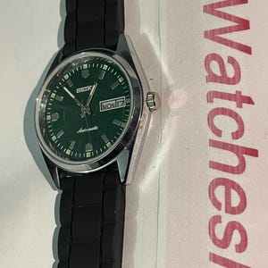 May include: A Seiko automatic wristwatch with a green dial and a black rubber strap. The watch face displays the day and date, with the day reading "MON 17". The watch has a silver-colored case.