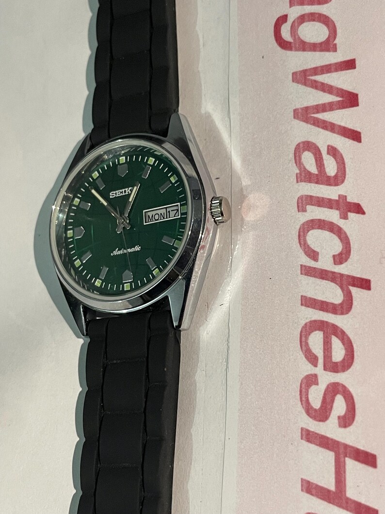 May include: A Seiko automatic wristwatch with a green dial and a black rubber strap. The watch face displays the day and date, with the day reading "MON 17". The watch has a silver-colored case.