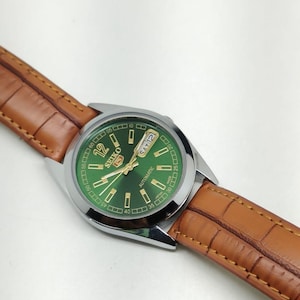 Peut inclure: Une montre automatique Seiko argentée avec un cadran vert et des détails dorés. La montre affiche le jour et la date et est montée sur un bracelet en cuir marron. Le cadran affiche le texte "SEIKO AUTOMATIC".