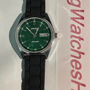 May include: A Seiko automatic wristwatch with a green dial and a black rubber strap. The watch face displays the day and date, with a silver-colored case. The watch has a grid pattern on the dial.