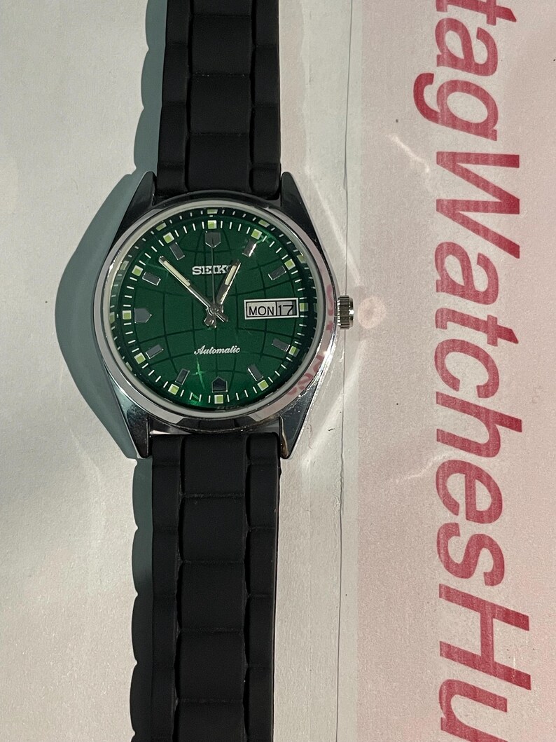 May include: A Seiko automatic wristwatch with a green dial and a black rubber strap. The watch face displays the day and date, with a silver-colored case. The watch has a grid pattern on the dial.
