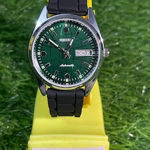 May include: A Seiko automatic wristwatch with a green dial and black rubber strap. The watch face displays the day and date. The watch is on a yellow display stand with the text "VintagWatchesHub".