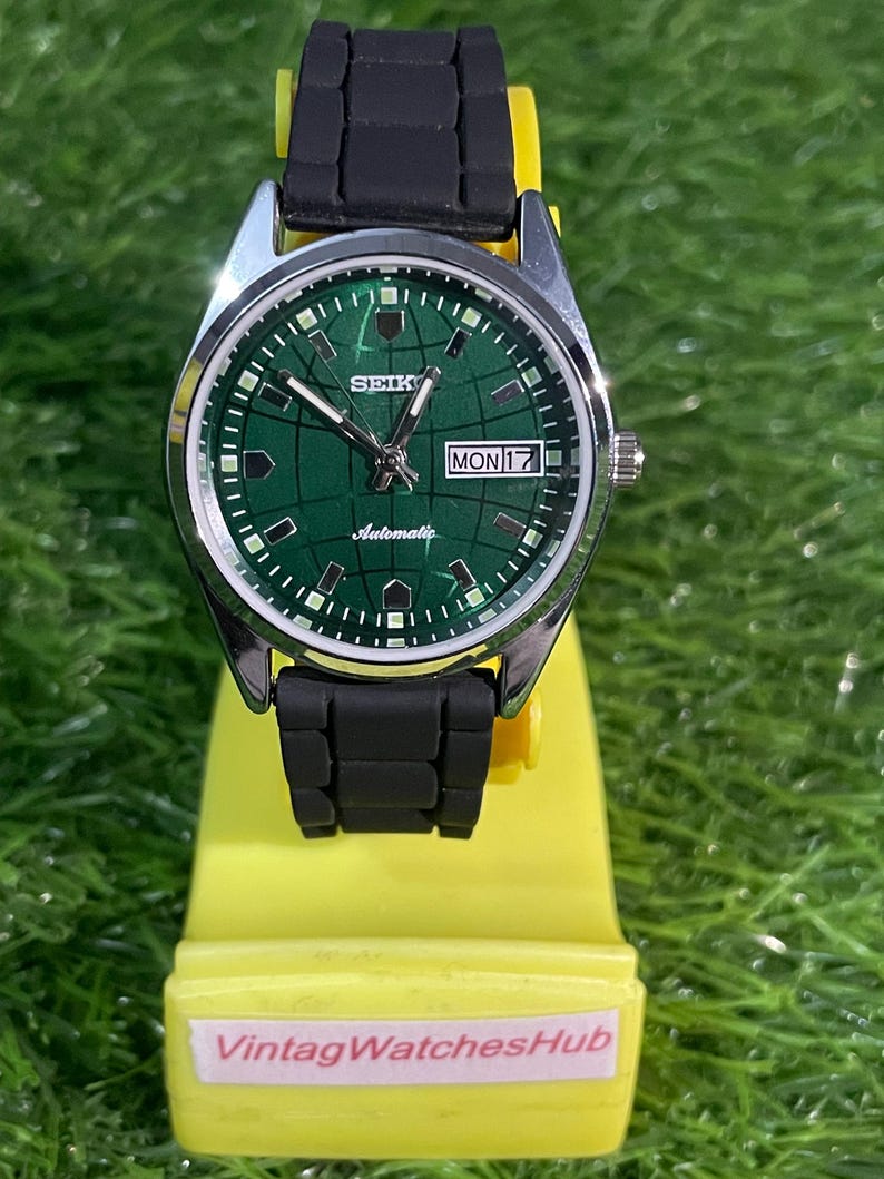 May include: A Seiko automatic wristwatch with a green dial and black rubber strap. The watch face displays the day and date. The watch is on a yellow display stand with the text "VintagWatchesHub".