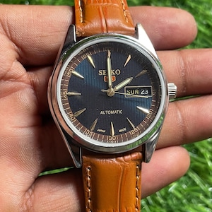 Op de afbeelding: Een Seiko 5 automatisch horloge met een zilveren kast, donkerblauwe wijzerplaat en bruine leren band. De wijzerplaat toont de dag en datum, met goudkleurige wijzers en markeringen. De band is lichtbruin.