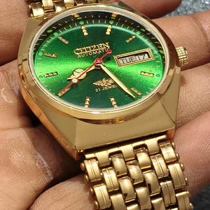 Può includere: Un orologio da polso automatico Citizen color oro con un quadrante verde brillante, indici dorati e una finestrella per giorno e data a ore tre. L'orologio è dotato di un cinturino a maglie in metallo color oro coordinato.