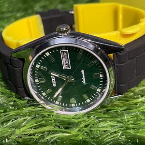 May include: A close-up of a Seiko automatic wristwatch with a dark green face and silver hands. The watch has a black rubber strap and a yellow watch holder. The watch face displays the day of the week and the word "Automatic".