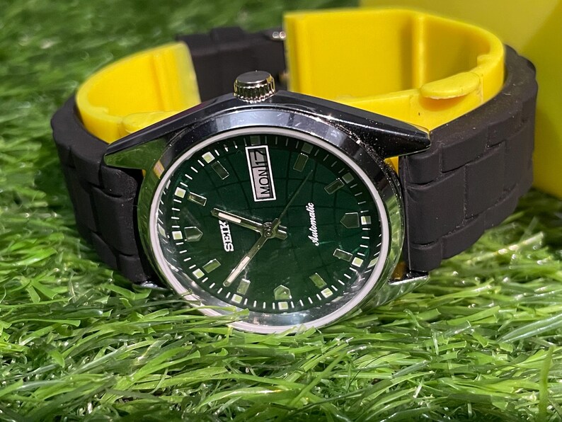 May include: A close-up of a Seiko automatic wristwatch with a dark green face and silver hands. The watch has a black rubber strap and a yellow watch holder. The watch face displays the day of the week and the word "Automatic".