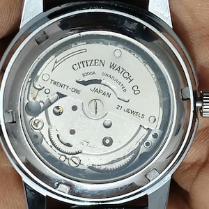 Pode incluir: Uma caixa de relógio prateada com movimento mecânico exposto. A placa interna apresenta o texto CITIZEN WATCH CO, B200A, UNADJUSTED, TWENTY-ONE, JAPAN e 21 JEWELS. O relógio tem um acabamento metálico e uma coroa de ajuste lateral.