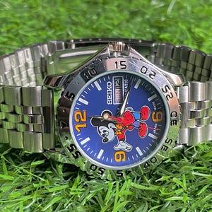 Op de afbeelding: Een Seiko polshorloge met een roestvrijstalen band en een blauwe wijzerplaat met een Mickey Mouse-afbeelding. Het horloge heeft een zilveren lunette met zwarte cijfers en een datumweergave. Het horloge ligt op groen gras.