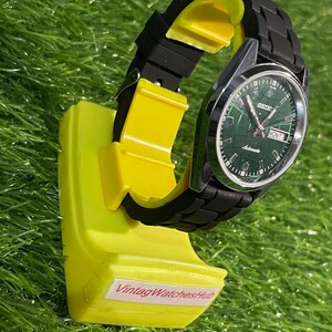 May include: A vintage Seiko automatic watch with a green dial and black band is displayed on a bright yellow stand. The watch face shows the day and date. The stand has the text "VintagWatchesHub".
