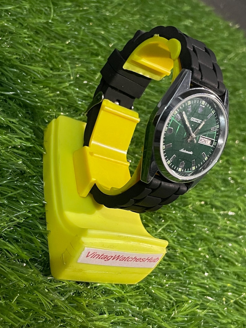 May include: A vintage Seiko automatic watch with a green dial and black band is displayed on a bright yellow stand. The watch face shows the day and date. The stand has the text "VintagWatchesHub".