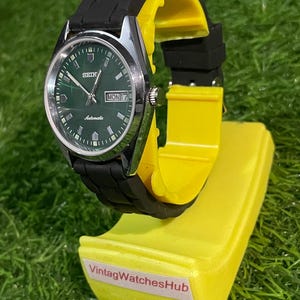 May include: A vintage Seiko automatic watch with a green dial and black rubber strap. The watch is displayed on a yellow stand with the text "VintagWatchesHub". The watch face shows the day and date.