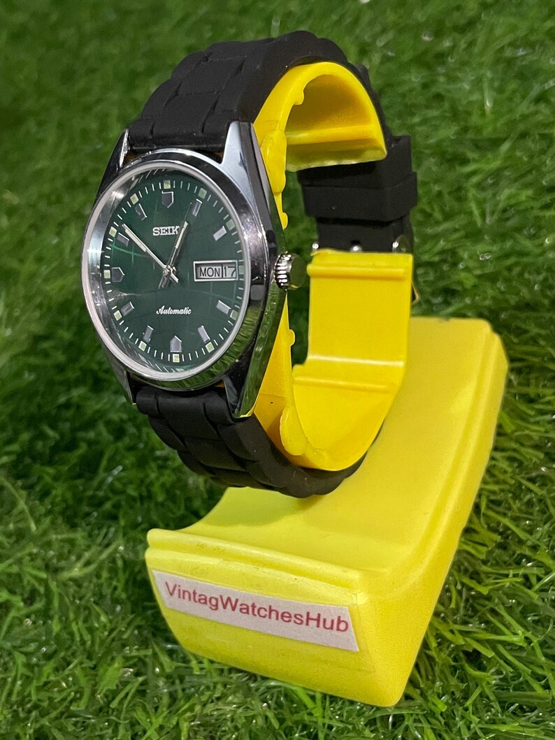 May include: A vintage Seiko automatic watch with a green dial and black rubber strap. The watch is displayed on a yellow stand with the text "VintagWatchesHub". The watch face shows the day and date.