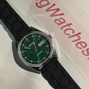 May include: A vintage Seiko automatic watch with a green dial and a black rubber strap. The watch face displays the day and date. The watch has a silver-colored case and a black textured band.