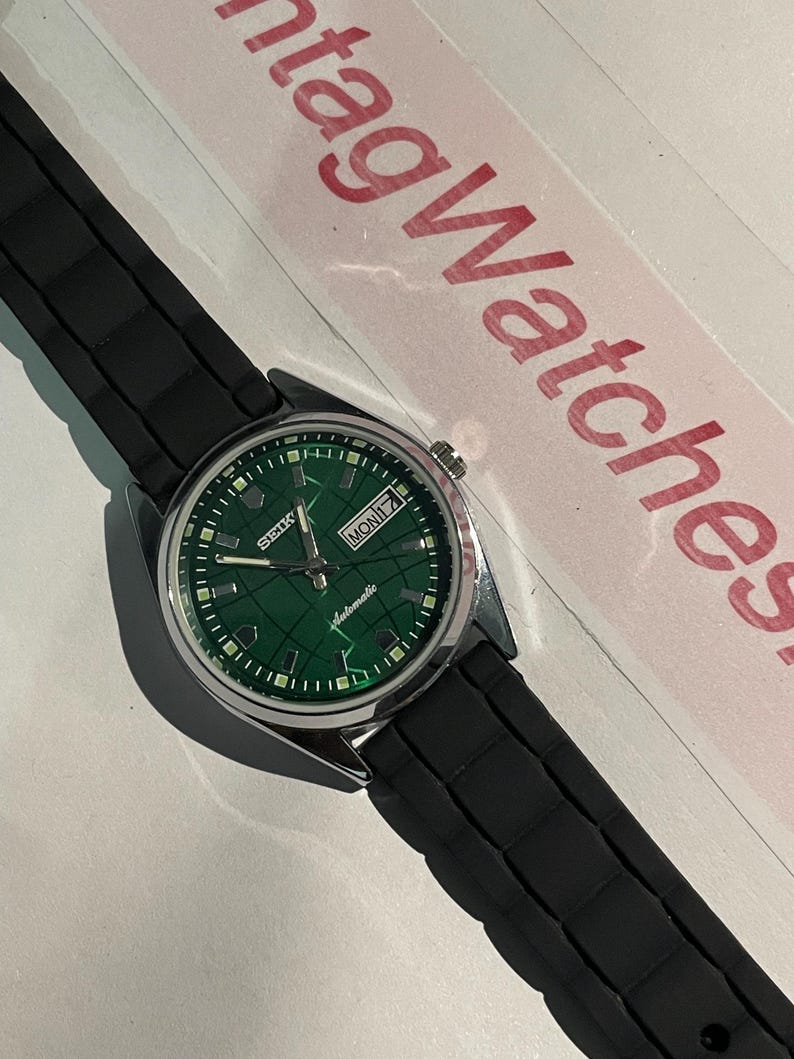 May include: A vintage Seiko automatic watch with a green dial and a black rubber strap. The watch face displays the day and date. The watch has a silver-colored case and a black textured band.
