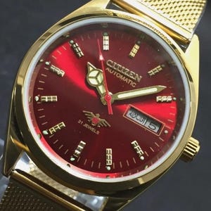 Puede incluir: Reloj Citizen Automatic dorado con esfera roja y agujas doradas. El reloj tiene una correa de malla y una pantalla de fecha. La esfera incluye el texto "CITIZEN AUTOMATIC" y "21 JEWELS".