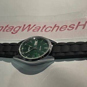 May include: A silver-toned wristwatch with a green dial and black band. The watch face displays the day and date. The band is made of black rubber. The text "VintagWatchesHu" is visible in the background.