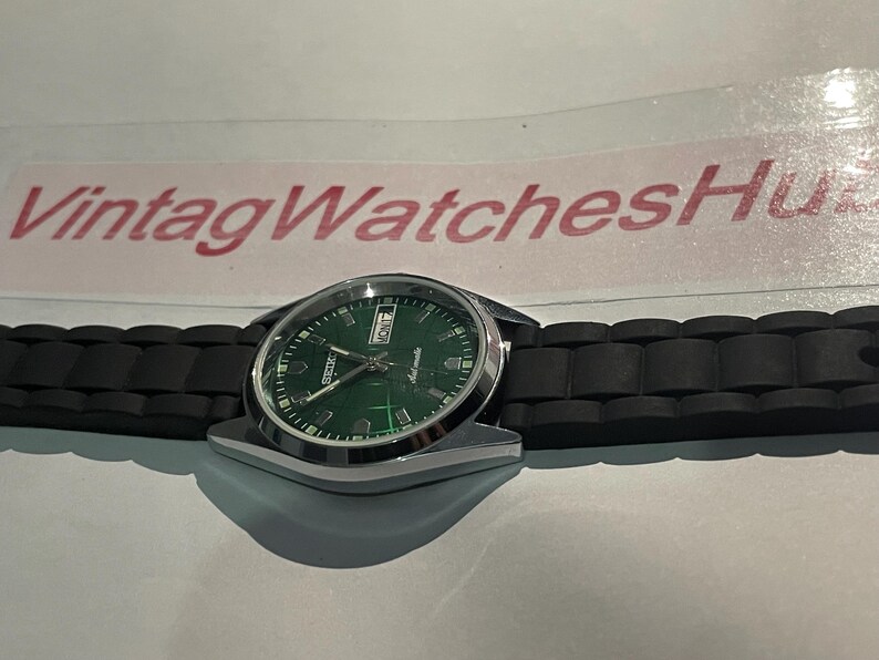 May include: A silver-toned wristwatch with a green dial and black band. The watch face displays the day and date. The band is made of black rubber. The text "VintagWatchesHu" is visible in the background.