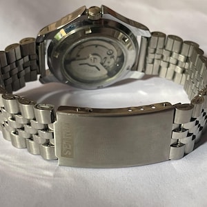 May include: A stainless steel wristwatch with a silver-toned band. The watch's back is visible, showcasing the inner workings. The band features a series of connected rectangular links. The clasp is engraved with the brand name "SEIKO".