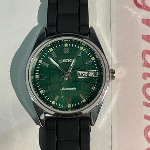 May include: A silver-toned Seiko wristwatch with a green face and black band. The watch face displays the day and date, with the word "Automatic" printed below the hands. The watch band is black, and the watch is set against a white background.