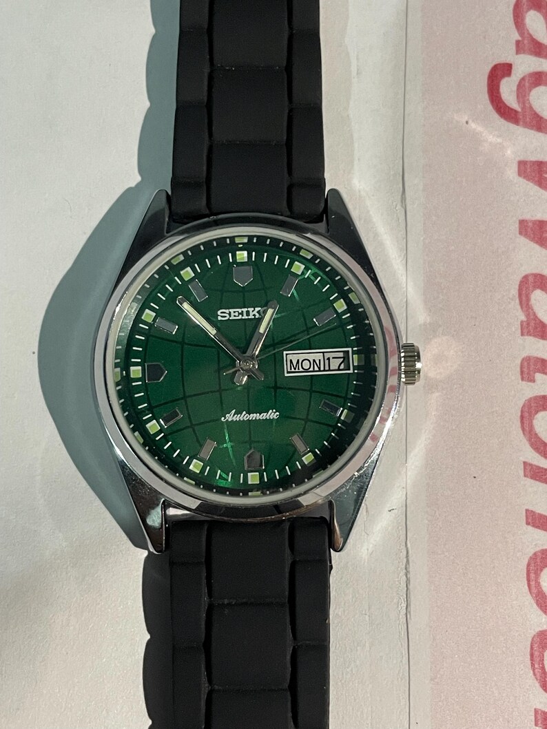 May include: A silver-toned Seiko wristwatch with a green face and black band. The watch face displays the day and date, with the word "Automatic" printed below the hands. The watch band is black, and the watch is set against a white background.