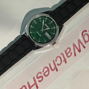 May include: A silver-toned wristwatch with a green dial and black rubber strap. The watch face displays the day and date, with the brand name "Seiko" visible. The watch is set against a white background.