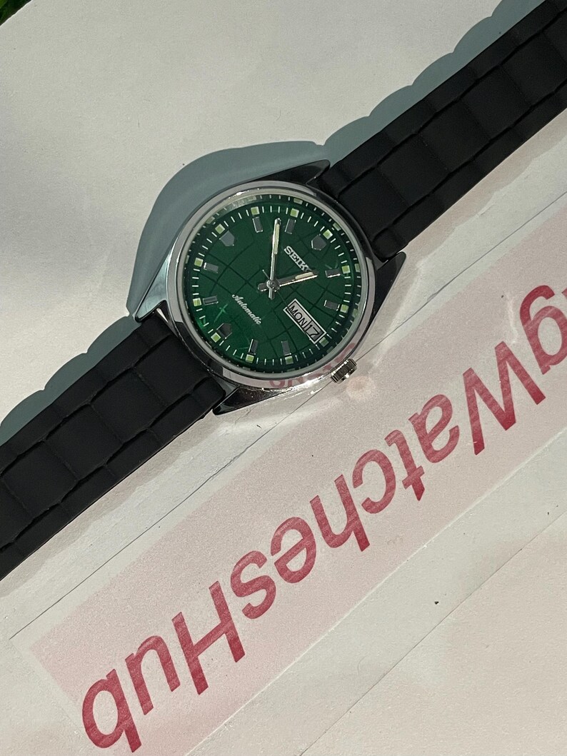 May include: A silver-toned wristwatch with a green dial and black rubber strap. The watch face displays the day and date, with the brand name "Seiko" visible. The watch is set against a white background.