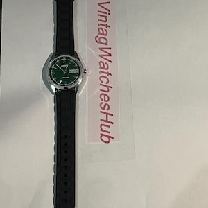 May include: A silver-toned wristwatch with a green dial and black rubber strap. The watch face displays the day and date. The strap is textured. The text "VintagWatchesHub" is visible on a pink label.