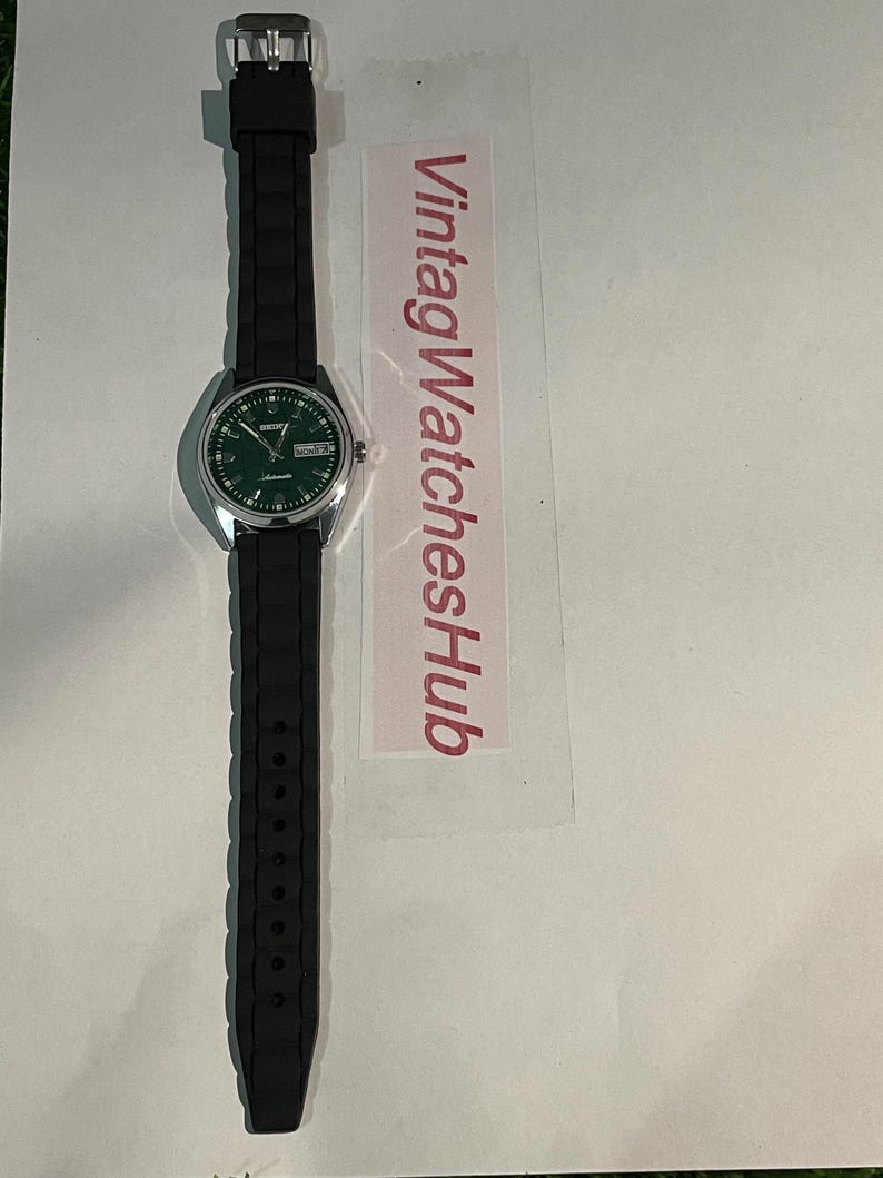 May include: A silver-toned wristwatch with a green dial and black rubber strap. The watch face displays the day and date. The strap is textured. The text "VintagWatchesHub" is visible on a pink label.