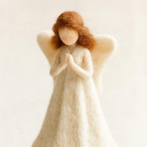 Puede incluir: Figura de ángel de fieltro hecha a mano con vestido y alas color crema y cabello castaño rojizo. El ángel está sobre una pequeña base redonda de madera. Las manos están juntas en oración.