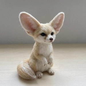 May include: A needle-felted fennec fox figurine featuring oversized, pointed ears with soft pink inner coloring, a cream-colored chest, and a light tan coat. The small sculpture has dark, expressive eyes, a tiny brown nose, and a bushy tail.