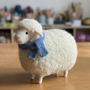 May include: A handcrafted, white sheep figurine with a blue knitted scarf. The sheep has a fluffy, textured wool body and small wooden legs. The figurine is displayed on a wooden surface, showcasing its charming details.