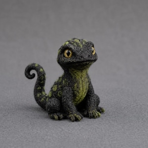 Può includere: Una statuetta di lucertola realizzata con la tecnica dell'infeltrimento ad ago, composta da fibre di lana grigio antracite e verde oliva. La creatura presenta un motivo maculato, una coda arricciata e grandi occhi ambrati con pupille nere.