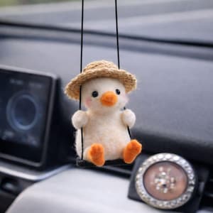 May include: A handmade, white felt duckling wearing a straw hat, suspended by black strings. The duckling has an orange beak and feet, and is designed to hang as a car accessory. The background is blurred, suggesting an interior car setting.