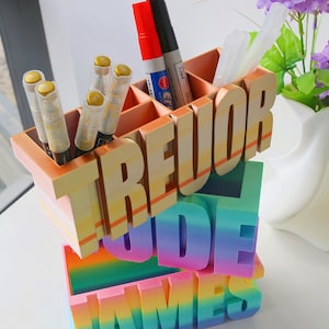 Pu&ograve; includere: Un organizer da scrivania a pi&ugrave; livelli con i nomi "TREVOR", "JUDE" e "JAMES" in grassetto e colorati. La sezione superiore contiene penne e pennarelli. Un accessorio da scrivania vivace e decorativo.