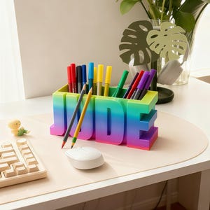 Pu&ograve; includere: Un colorato organizer da scrivania a forma del nome "JUDE" contiene penne e matite. L'organizer ha una sfumatura di colori, che va dal blu al viola. Un mouse e una tastiera per computer bianchi sono in primo piano.